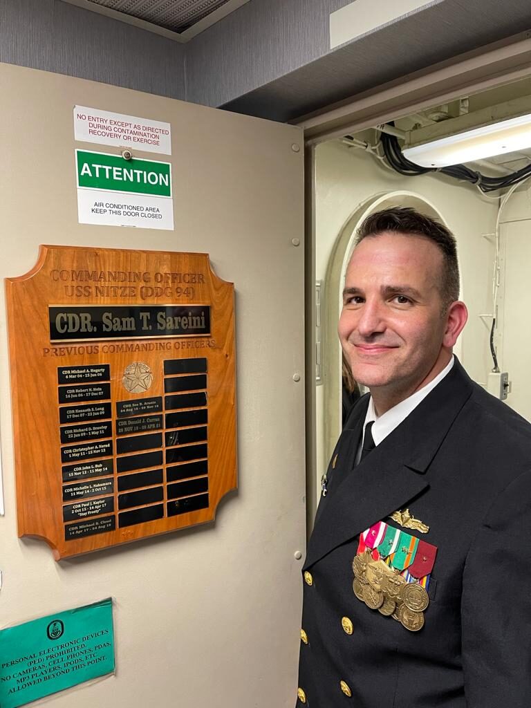Commanding Officer Sam Sareini standing in front of a plaque with his name as a commander of the U.S. naval ship.